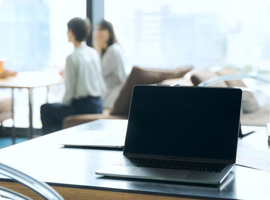 デスク上のノートPCと奥に談笑する2人のビジネスパーソンの写真